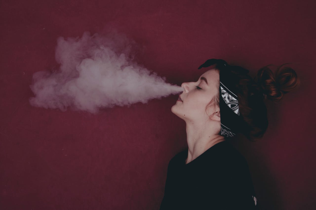 Young woman vaping, releasing a cloud of smoke against a red background.